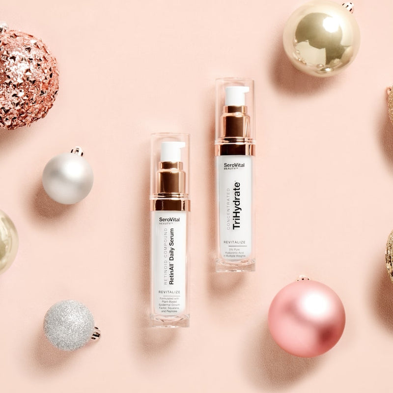 A bottle of anti-wrinkle serum RetinAll Daily Serum next to a bottle of hyaluronic acid formula TriHydrate Concentrate on a pink background with holiday ornaments.